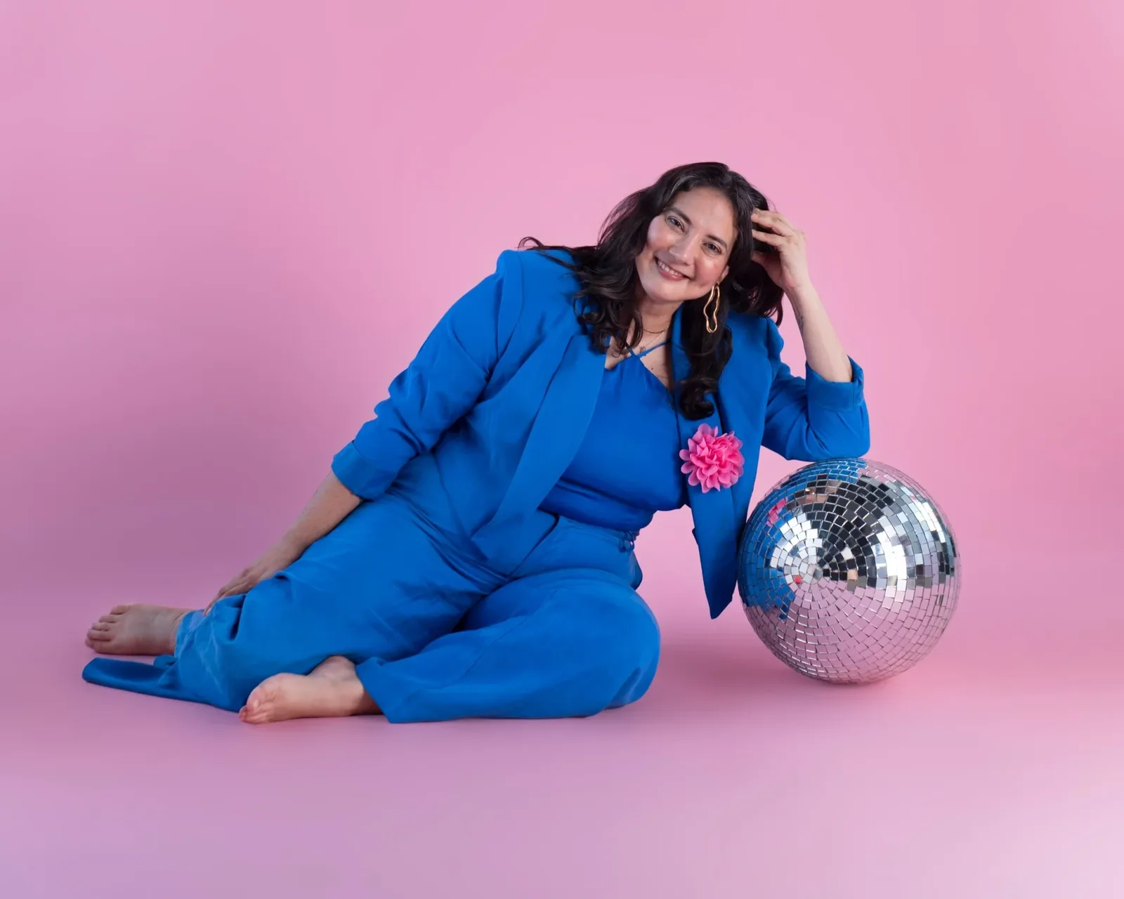 Alejandra Porta in a bright blue suit, leaning on a mirrored disco ball against a pink backdrop